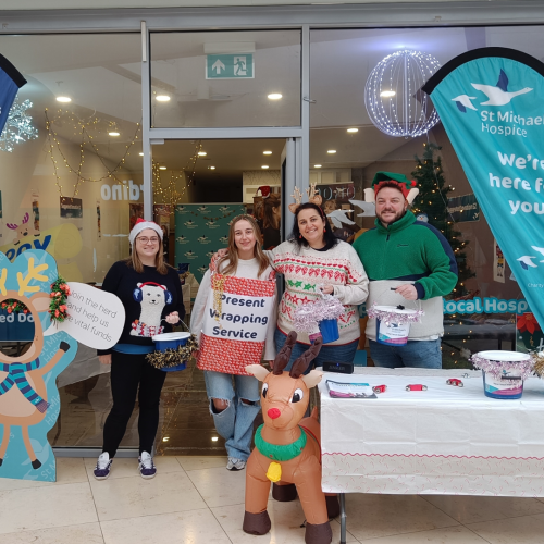 Four people wearing Christmas jumpers outside a shop, holding collection buckets and smiling at the camera. 