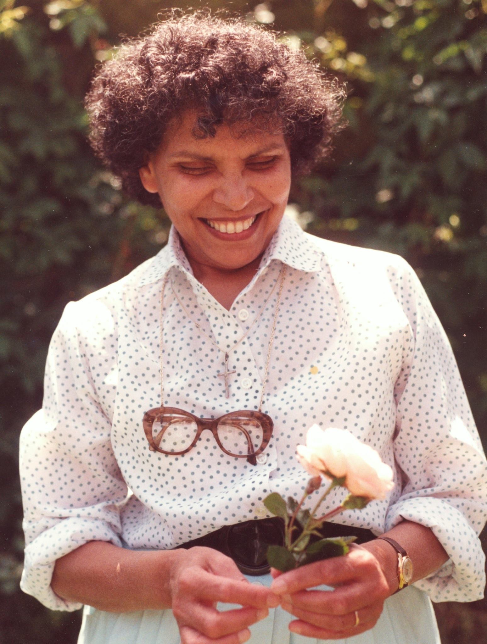 Photograph of Iris wearing a white polka dot shirt, smiling and holding flowers.